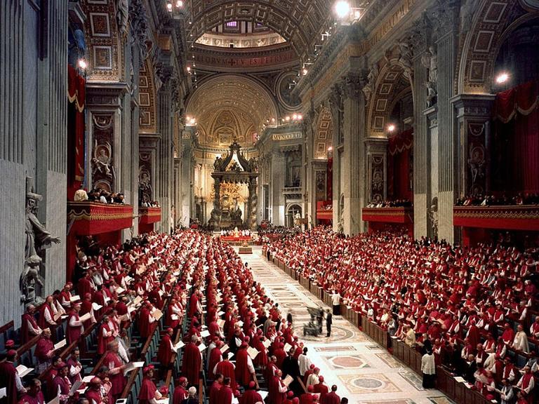 Bischöfe tagen in der Konzilsaula in Rom. Alle tragen Gewänder. Es sind etwa 2.800 so genannte Konzilsväter, sie sitzen einander in zwei gegenüberliegenden Bankreihen gegenüber.